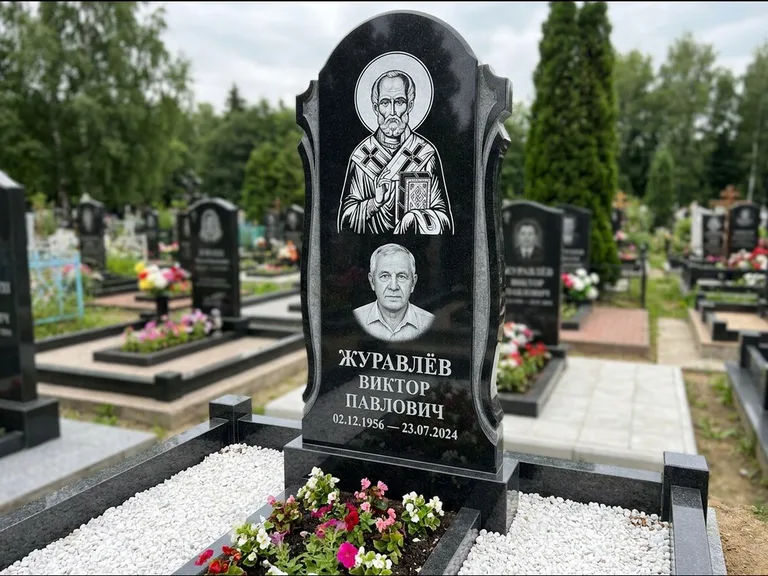 Памятник с гравировкой иконы: стела с образом в полный рост на обороте — гравировка образа Матроны Московской, низкие кусты спиреи в цвету, работа «Лебедева» в Саратове